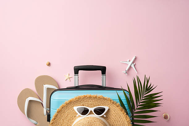 Travel essentials arranged on a pastel pink background creating a summer vibe. Vacation preparation theme with a suitcase, hat, and flip-flops complemented by tropical accents for a sunny adventure