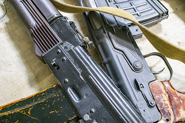 Collection of firearms on a neutral surface, with two distinct rifles matte black, ribbed grip panels darker, textured grip, synthetic stock and an old shotgun in the background Soft, diffuse lig
