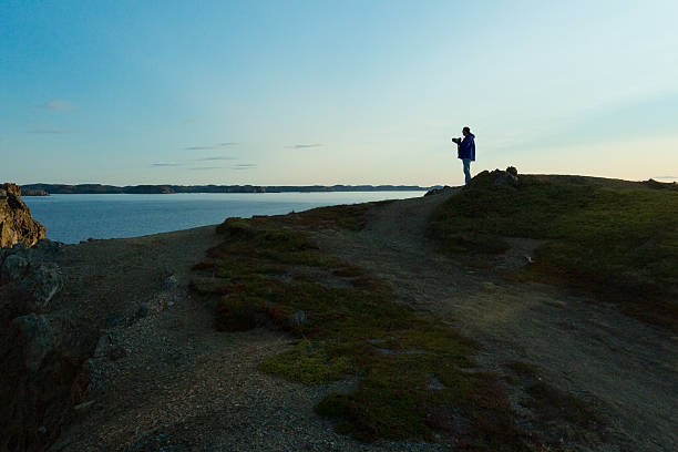 A male photographer stands looking over a body of water from a rocky cliff. The daylight is waning into golden hour.