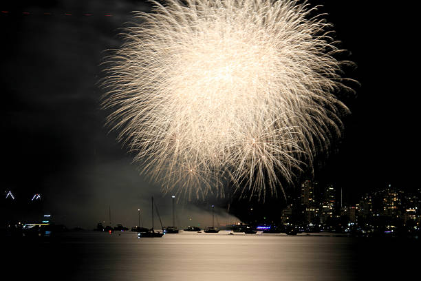 A vibrant display of fireworks illuminates the night sky above English Bay in Vancouver, British Columbia. The colorful explosions reflect over the calm ocean water