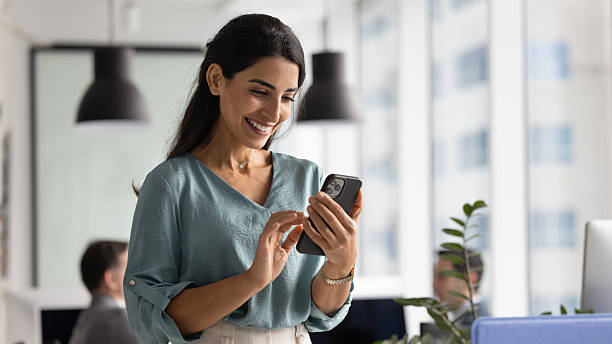 Happy beautiful Hispanic businesswoman using web service on smartphone in office workspace, working on online project on mobile phone, typing on cellphone, smiling. Banner shot