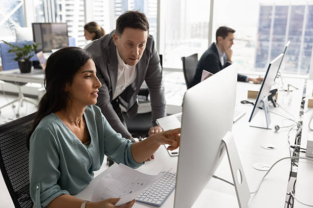 Boss and employee discussing work task at computer monitor in open office space. Female Latin professional showing online content, work report to male team leader at workplace