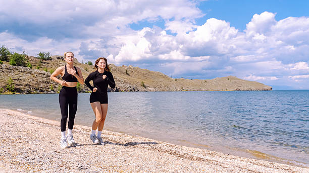 Two women jog along a beautiful beach, focusing on their cardio workout under the sunny sky.