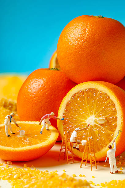 istock Miniature Workers Painting a Giant Orange Slice. Workers decorating orange fruit with gold seed on blue background 2227314421