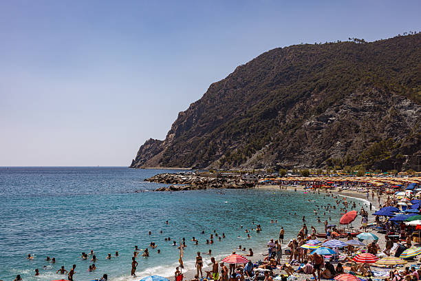 An energetic midsummer day at Monterosso al Mare in Cinque Terre, Italy, captures a crowded beach scene with colorful umbrellas and throngs of tourists enjoying the clear blue Mediterranean waters. The dramatic rocky cliffs serve as a scenic backdrop, highlighting the vibrant atmosphere, popular vacation spirit, and iconic beauty of Italy’s Ligurian coastline. Perfect for themes about Mediterranean travel, European summer holidays, beach destinations, and leisure culture.