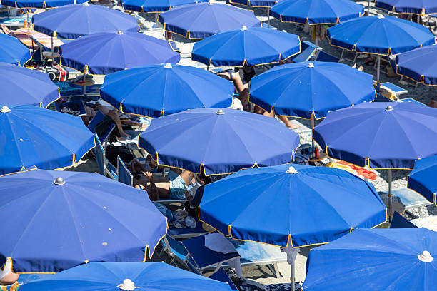 A lively summer scene at Monterosso al Mare in Cinque Terre, Italy, features vibrant blue umbrellas packed closely together on the sandy beach. Holiday-goers relax beneath the shade, enjoying the warm Mediterranean sun and lively atmosphere. The image captures the festive spirit, relaxation, and scenic beauty that make Monterosso one of the most popular beach destinations on the Italian Riviera, perfect for illustrating summer vacations, Mediterranean lifestyle, and European seaside escapes.
