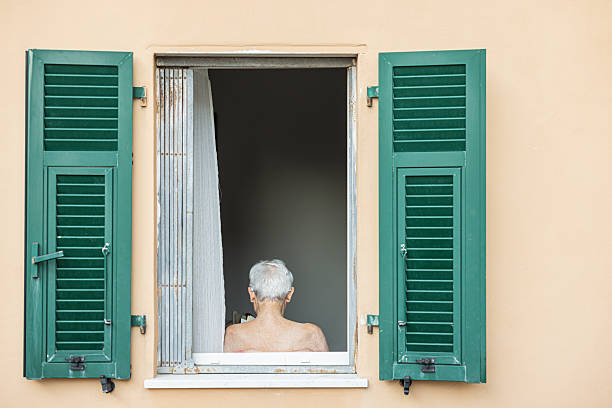 An elderly man with gray hair is seen from behind, gazing out of an open window framed by pastel peach walls and traditional green shutters. The natural light highlights the peaceful solitude of an interior space, suggesting themes of contemplation, senior living, and the timeless beauty of everyday moments in an Italian or Mediterranean setting. The simple yet evocative scene is perfect for concepts relating to aging, peaceful mornings, domestic life, relaxation, and the charm of classic European architecture.