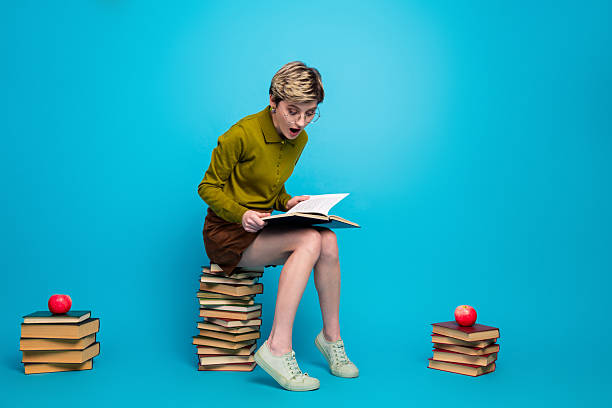 istock Charming young woman with short hair reading a book while sitting on stacked book against a vivid blue background 2227327708