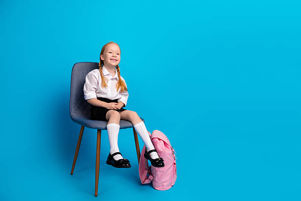 Adorable young schoolgirl wearing a uniform and sitting on a chair against a blue background with a pink backpack nearby