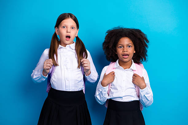 istock Charming schoolgirls in uniforms surprised while standing against blue background, showcasing curiosity and friendship 2227334796