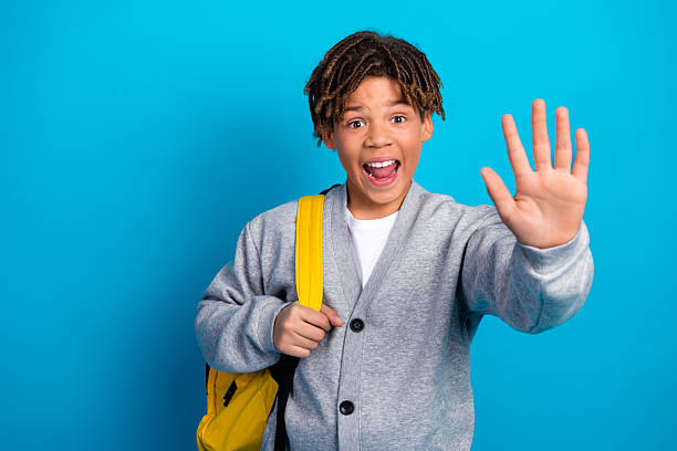 istock Excited young schoolboy with a backpack making a friendly hand gesture against a bright blue background 2227335293