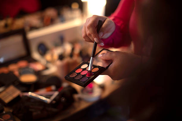 A close-up of hands using a makeup brush to mix colors on a vibrant eyeshadow palette in a studio environment, with blurred cosmetics in the background