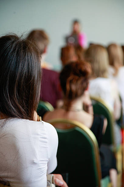 A rear-view shot captures a diverse group of unrecognizable individuals seated in rows, attentively listening to a speaker or presentation (blurred in the background) at a seminar, conference, or educational event. The focus on the audience's backs creates a sense of engagement and participation in a learning or professional environment.