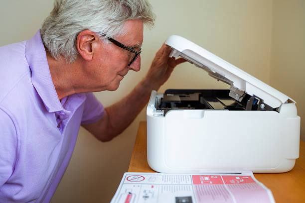 A senior man installing a new white inkjet printer in his home office. He refers to the instruction manual for assistance.