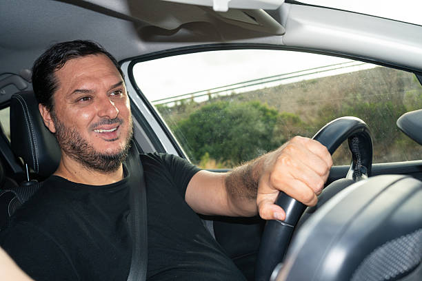 Latin man is happily driving a car along a winding country road. He shows excitement while navigating. The overcast sky hints at possible rain, creating a calm driving atmosphere.