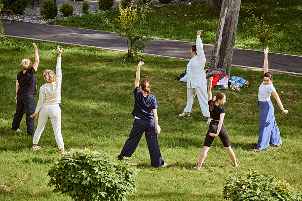 Adults, diverse ethnicities, practice qigong on grass under sunny skies. Instructed by mature woman in white, serene setting enhances focus and harmony