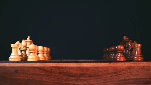 Lined up off white cream colored and dark brown wood chess pieces on checked pattern wooden chess board for concept of traditional war, battle, face off, confrontation between enemies stage theatre scene against black background with copy space