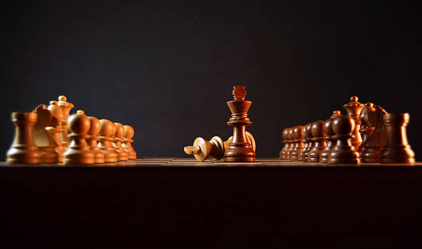 Off white king falling defeated and brown king winning -  Lined up off white cream colored and dark brown wood chess pieces on wooden board for concept of defeat, failure, victory, win traditional war, battle, face off, confrontation between enemies stage theatre scene against black background with copy space