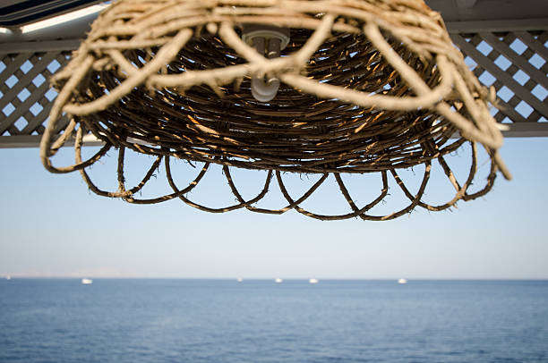 Wicker lampshade suspended from pergola frames calm ocean view. Bright sunlight highlights intricate weave against clear blue sky. Horizon dotted with distant boats