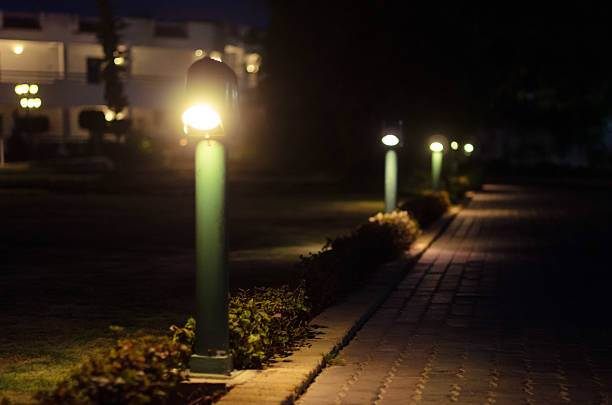 Warm lighting casts soft glow along hotel garden pathway at night. Neatly aligned lamps create serene ambiance with nearby foliage enhancing scene. Distant building is softly visible