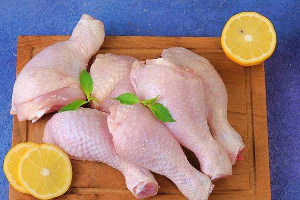Top angle of raw chicken legs with a halved lemon on wooden cutting board over blue surface.