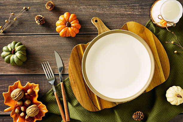 istock Rustic autumn table setting with white plate on pumpkin-shaped cutting board, green cloth, orange bowl of nuts, decorative pumpkins, pinecones, and candle. Cozy fall composition perfect for Thanksgiving or seasonal dining. 2227368840