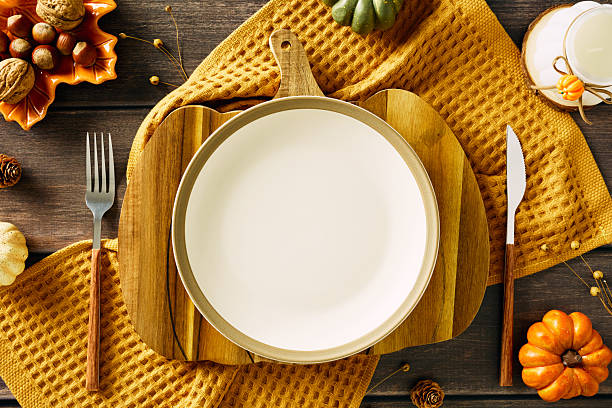 istock Warm autumn table setting with white plate on cutting board, yellow cloth, rustic cutlery, small pumpkin, pinecones, nuts, and ceramic candle. Cozy flat lay ideal for Thanksgiving 2227368907