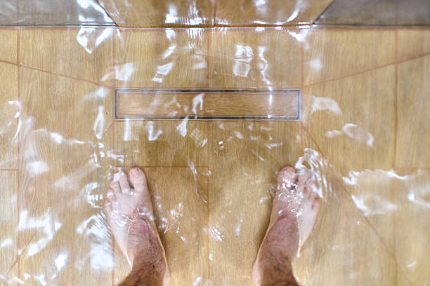 Overhead view of standing water on shower floor due to clogged drain. Tiled surface with visible water ripples enhances sense of disruption. Natural lighting captures scene in residential setting