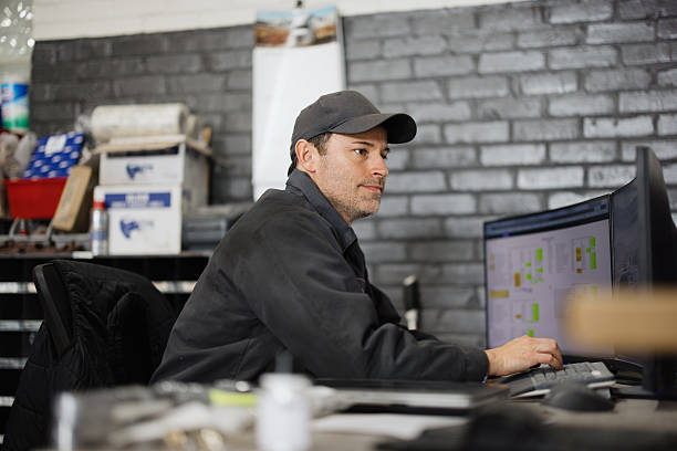 Mid adult male mechanic sitting at a desk, using computer software for checking car parts inventory and reviewing schematics
