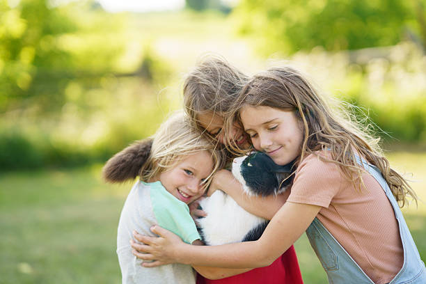 Three joyful children embrace a cat, sharing a heartfelt moment outdoors in a sunlit and lush countryside.