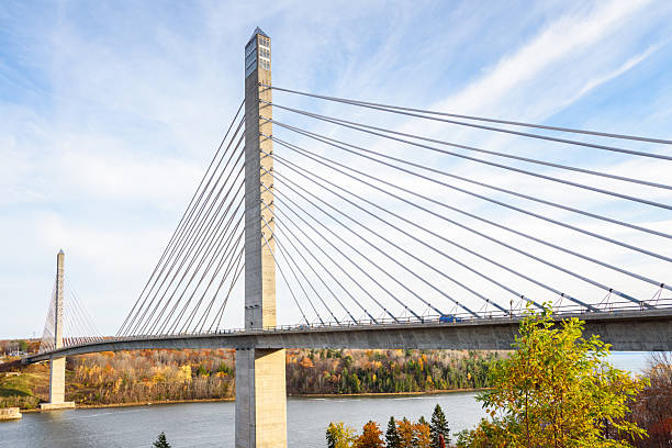 Modern suspension road bridge supported by concrete pylons spanning a river with forested banks in Maine on sunny autumn day. Penobscot, ME, USA.