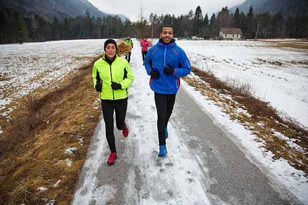 A happy and determined multi-racial couple in front, dressed in vibrant winter running gear (a lime green jacket for one, a blue jacket for the other), are jogging together on a partially snow-covered asphalt road. The background features a serene winter landscape with snow-dusted fields, a dark forest, anothe running couple and mountains, emphasizing outdoor fitness and shared perseverance in cold weather.