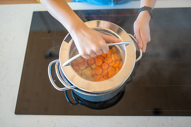 A multiracial woman in a brightly lit modern kitchen stirs simmering carrots in a pot, preparing homemade baby food with care and focus.