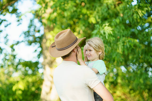 A man wearing a hat interacts sweetly with a child in a lush green outdoor setting, highlighting familial love and a warm, care-free summertime mood.
