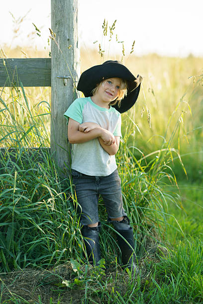 A cheerful child wearing a wide-brimmed hat posing by a wooden post in a vibrant grassy field under warm sunlight, exuding a playful and carefree countryside vibe.