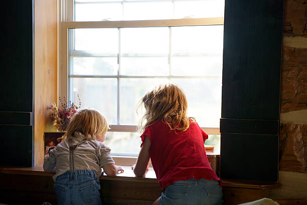 istock Two Children Curiously Looking Out of a Bright Window Inside a Rustic Room 2227406200