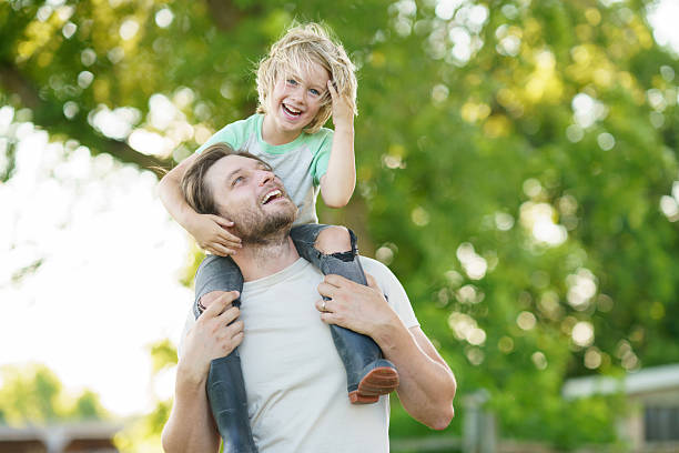 A joyful father carrying his cheerful young son on his shoulders outside during a sunny day surrounded by greenery, expressing family bonding and happiness.