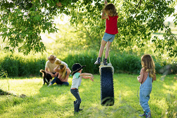 Several children enjoying a sunny afternoon playing with a rope swing on a farm, surrounded by lush greenery, while a family with a dog relaxes in the distance.