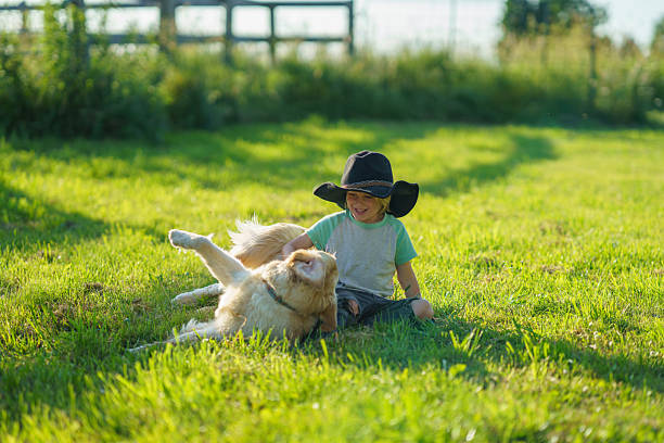 A cheerful boy wearing a hat enjoys time with his playful dog in a sunlit yard, surrounded by the beauty of nature.