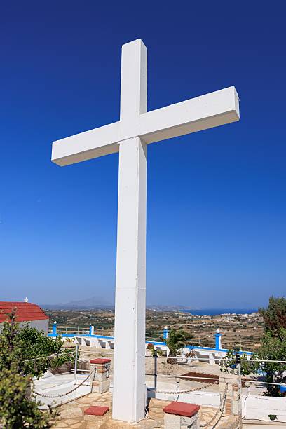 A tall white cross at a hilltop chapel in Kos, Greece, standing under a clear summer sky with views of the surrounding landscape.