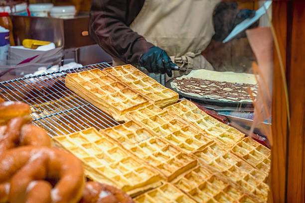 Golden waffles cooling on metal rack while cook prepares crepe with chocolate at food stand. Concept of fresh baking, sweet indulgence, street market cooking