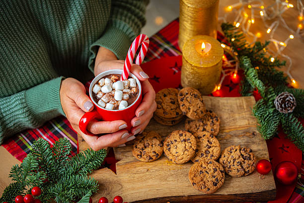 istock Woman warming her hands with a hot chocolate mug on cozy table with chocolate chips cookies, burning candles and ornaments 2227418382