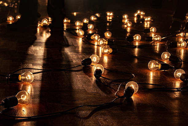 Multiple glowing vintage light bulbs scattered across a dark wooden floor, with human silhouettes and shadows visible in the background. The ambient scene features low lighting and a moody indoor atmosphere. No faces or identifying features are visible.