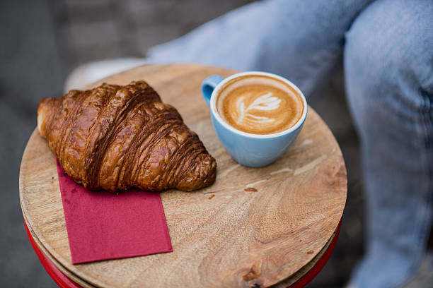 A close-up shot of a small outdoor table featuring a cappuccino with milk froth art and a flaky croissant, set for a peaceful park break.
