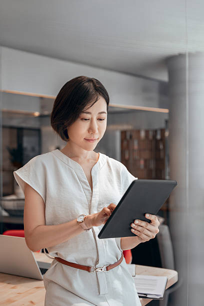 istock Middle-aged eastern businesswoman holding digital tablet using tab application standing at workplace in office. Mature Asian professional CEO business woman working on touchscreen computer. Vertical 2227434545