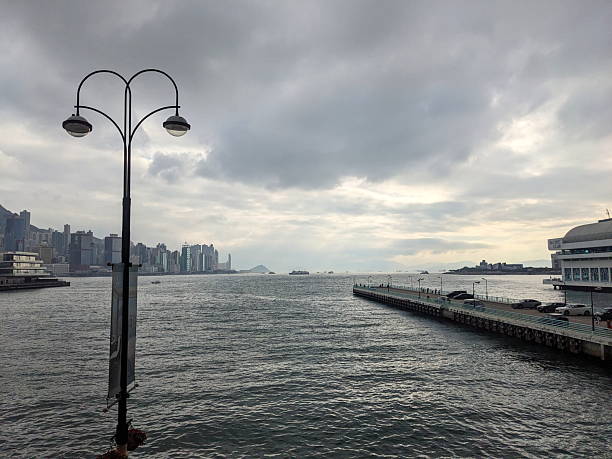 Hong Kong - June 01 2025: A view across Victoria Harbour towards the Hong Kong Island skyline under a dramatic, overcast sky, with a pier and waterfront promenade in the foreground