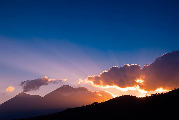 Sunlight bursts from behind volcanic peaks, casting golden rays across fluffy clouds and a deep blue sky. A peaceful sunrise in a natural landscape filled with awe and tranquil energy.