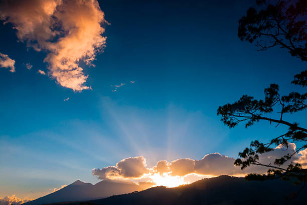 Sunlight bursts from behind volcanic peaks, casting golden rays across fluffy clouds and a deep blue sky. A peaceful sunrise in a natural landscape filled with awe and tranquil energy.