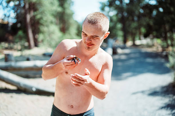 A smiling teenage boy with Down syndrome stands on the river's edge and looks at the crawdad he caught while swimming at the river with his family on a hot summer afternoon in Central Oregon.