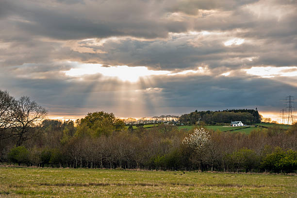Scenic rural landscape with crepuscular rays of sunlight streaming through dramatic clouds at sunset. Rolling fields, trees, and a farmhouse create a peaceful countryside view